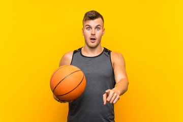 Young handsome blonde man holding a basket ball over isolated yellow background surprised and pointing front