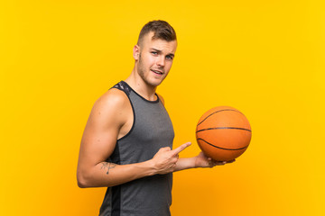 Fototapeta premium Young handsome blonde man holding a basket ball over isolated yellow background and pointing it
