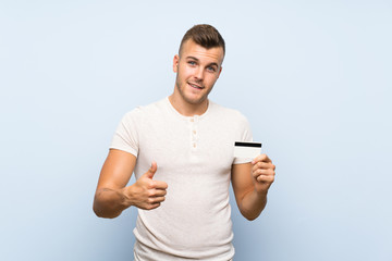Young handsome blonde man over isolated blue background holding a credit card