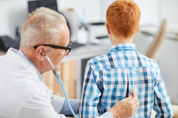 Back view of senior doctor using stethoscope listening to heartbeat and breathing of cute red haired boy during consultation in child healthcare clinic, copy space