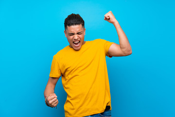 Young man with yellow shirt over isolated blue background celebrating a victory