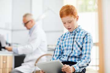 Portrait of cute red haired boy using digital tablet in waiting room at doctors office, copy space