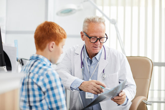 Portrait of white-haired senior doctor pointing at x ray image while talking to little patient during consultation in child healthcare clinic, copy space