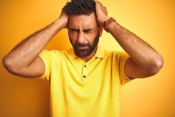 Young indian man wearing polo standing over isolated yellow background suffering from headache desperate and stressed because pain and migraine. Hands on head.
