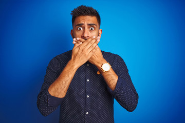 Young indian elegant man wearing shirt standing over isolated blue background shocked covering mouth with hands for mistake. Secret concept.