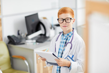 Portrait of cute red haired boy posing as doctor wearing white coat and smiling happily at camera holding clipboard, copy space