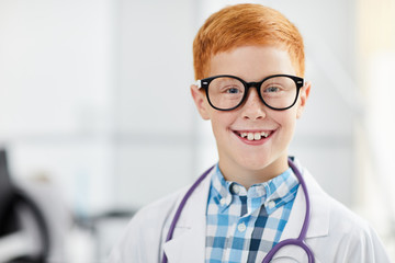 Head and shoulders portrait of freckled teenage boy posing as doctor wearing white coat and smiling happily looking at camera, copy space