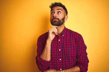 Young indian man wearing casual shirt standing over isolated yellow background with hand on chin thinking about question, pensive expression. Smiling with thoughtful face. Doubt concept.