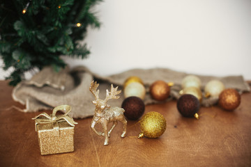 Obraz na płótnie Canvas Golden reindeer, gift box and glitter bauble toys on background of christmas tree with lights on rustic background. Merry Christmas and Happy Holidays. Festive decorations