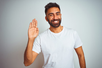 Young indian man wearing t-shirt standing over isolated white background Waiving saying hello happy...