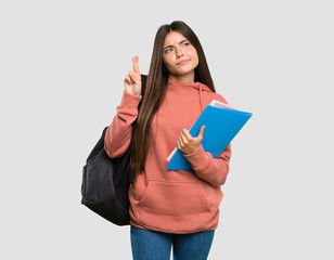 Young student woman holding notebooks with fingers crossing and wishing the best over isolated grey background