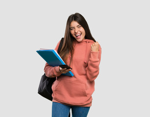Young student woman holding notebooks with phone in victory position over isolated grey background