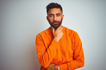 Young indian man wearing orange sweater over isolated white background looking confident at the camera with smile with crossed arms and hand raised on chin. Thinking positive.