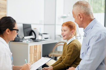 Fototapeta premium Side view portrait of smiling boy visiting doctor with father and looking at nurse filling patients form in waiting room, copy space