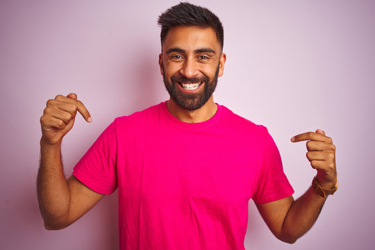 Young indian man wearing t-shirt standing over isolated pink background looking confident with smile on face, pointing oneself with fingers proud and happy.