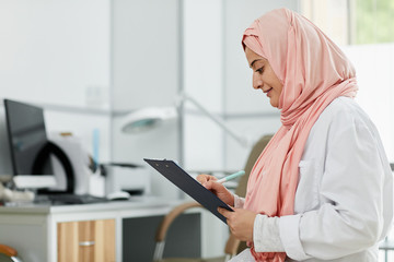 Side view portrait of Middle-Eastern woman wearing hijab working as nurse in medical clinic and writing on clipboard, copy space - obrazy, fototapety, plakaty