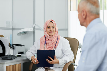 Portrait of young Arab woman working as nurse in medical clinic and talking to senior patient, copy...