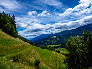 Obraz premium A stunning view on Alpine valley. Hiking trails in the high mountain. Green grass and bushes cover the slopes. A remote view on small settlement in the bottom of the valley. Spring in the mountains.