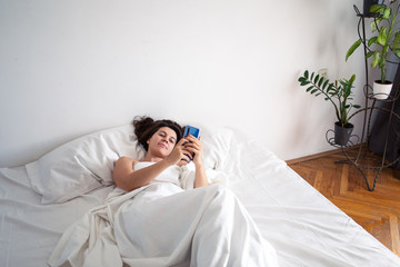 woman with smartphone in bed with white sheets