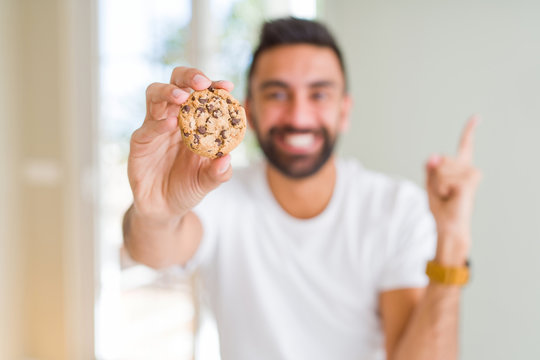 Handsome hispanic man eating chocolate chips cookies very happy pointing with hand and finger to the side