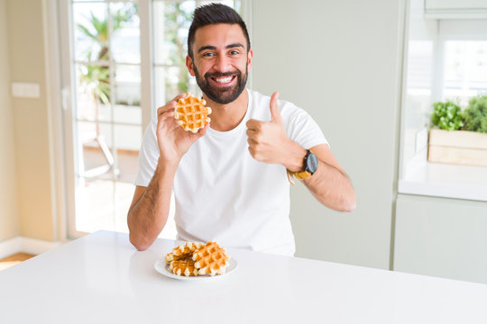 Handsome hispanic man eating sweet belgian waffle pastry happy with big smile doing ok sign, thumb up with fingers, excellent sign