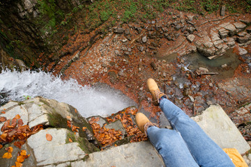 woman in blue jeans and brown boots sitting on the edge looking at waterfall
