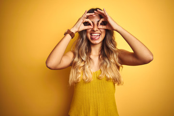 Young beautiful woman wearing t-shirt over yellow isolated background doing ok gesture like binoculars sticking tongue out, eyes looking through fingers. Crazy expression.