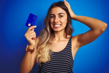 Fototapeta na wymiar Young beautiful woman holding credit card over blue isolated background stressed with hand on head, shocked with shame and surprise face, angry and frustrated. Fear and upset for mistake.
