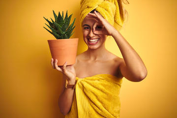 Young beautiful woman wearing a towel holding aloe vera pot over yellow isolated background with happy face smiling doing ok sign with hand on eye looking through fingers