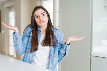 Beautiful young woman sitting on white table at home clueless and confused expression with arms and...