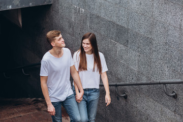 Obraz na płótnie Canvas Happy young couple walks the streets of the city and hold hands. guy and girl in white t-shirts and jeans outdoors. Teenagers hug on the background of the urban landscape. Couple close up portrait.
