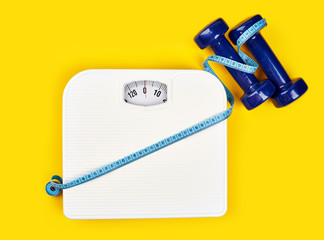 bathroom scales and measuring tape for weight loss concept on yellow background. Sport and diet for losing weight. Bathroom scale, measuring tape and dumbbell.