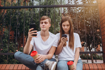 A young couple drinks coffee from cardboard cups and looks at smartphones. Teenagers guy and girl are sitting outdoors. Modern technologies. The problem of communication in modern society.