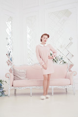 Obraz na płótnie Canvas young beautiful caucasian woman posing in pink retro dress indoors alone. slim adult girl in vintage outfit with old sofa on white background.