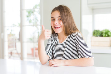 Beautiful young girl kid wearing stripes t-shirt doing happy thumbs up gesture with hand. Approving expression looking at the camera showing success.
