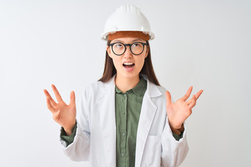 Young chinese engineer woman wearing coat helmet glasses over isolated white background crazy and mad shouting and yelling with aggressive expression and arms raised. Frustration concept.