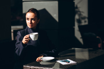 beautiful girl in a black coat sits on a summer terrace of a restaurant with a cup of coffee. Portrait of a young girl with a hard light. Business lady is drinking coffee in a street cafe.