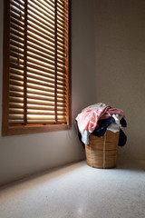 Heap of dirty clothes in wicker basket at corner in room near window are waiting to clean.