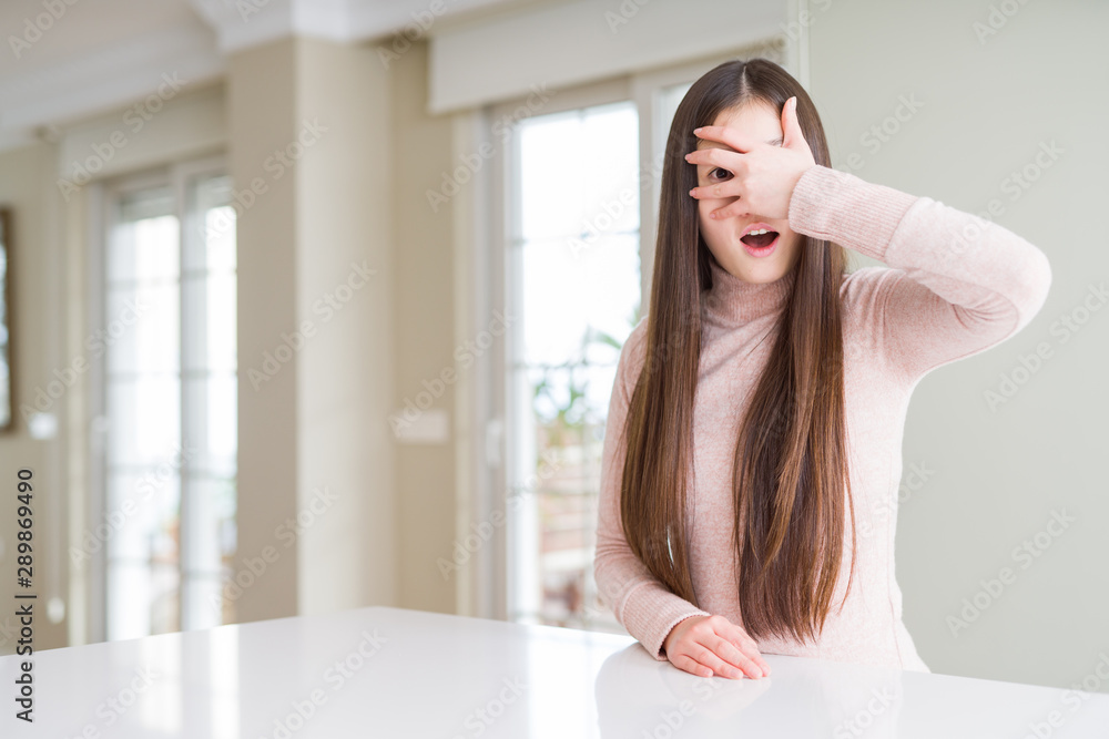 Poster Beautiful Asian woman wearing casual sweater on white table peeking in shock covering face and eyes with hand, looking through fingers with embarrassed expression.