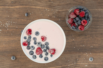 Yogurt smoothie with frozen raspberries, blueberries and coconut shavings over wooden background. Healthy food concept. Top view.