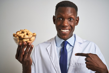 African american doctor man holding bowl with peanuts over isolated white background with surprise face pointing finger to himself