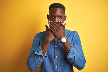Young african american man wearing denim shirt standing over isolated yellow background shocked covering mouth with hands for mistake. Secret concept.
