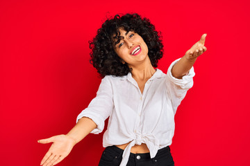 Young arab woman with curly hair wearing white casual shirt over isolated red background looking at...