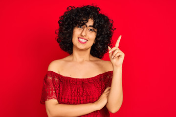 Young arab woman with curly hair wearing casual dress over isolated red background with a big smile on face, pointing with hand and finger to the side looking at the camera.