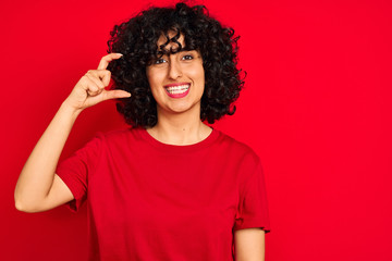 Young arab woman with curly hair wearing casual t-shirt over isolated red background smiling and confident gesturing with hand doing small size sign with fingers looking and the camera.