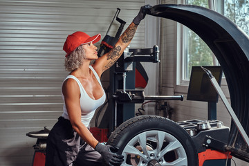 Obraz na płótnie Canvas Focused blond woman is doing man's job - she is fixing broken car at workshop.