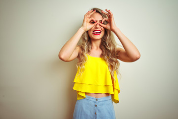 Obraz na płótnie Canvas Young beautiful woman wearing yellow t-shirt standing over white isolated background doing ok gesture like binoculars sticking tongue out, eyes looking through fingers. Crazy expression.