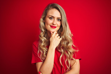 Young beautiful woman wearing basic t-shirt standing over red isolated background looking confident at the camera smiling with crossed arms and hand raised on chin. Thinking positive.
