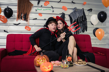 Halloween. young scared caucasian couple in vampires and witch clothing enjoying watching horror movie in television and sitting on sofa in halloween party at home, holiday, halloween festival concept