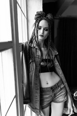 A beautiful young girl with an afro cornrows hairstyle, wearing a casual denim jacket and shorts, relaxes by the window and looks outside thoughtfully. Monochrome, black an white, fashion design.